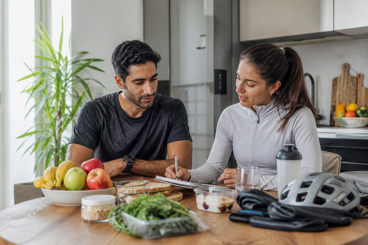 Very lean Middle Eastern endurance athlete around age 30 discussing a fueling plan with a female sports dietitian at a kitchen table, groceries and training gear nearby, bright practical editorial photography.