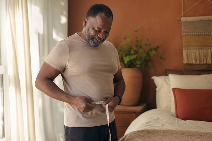 Black man around age 55 smiling subtly while measuring his waist in a sunlit bedroom after a long-term health change, relaxed realistic body, warm terracotta and cream lifestyle photography.
