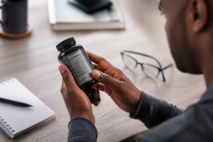 Close-up of a dark-skinned male hand carefully rotating an unbranded supplement bottle to inspect an intentionally unreadable facts panel, neutral desk and reading glasses, editorial product-detail photography.